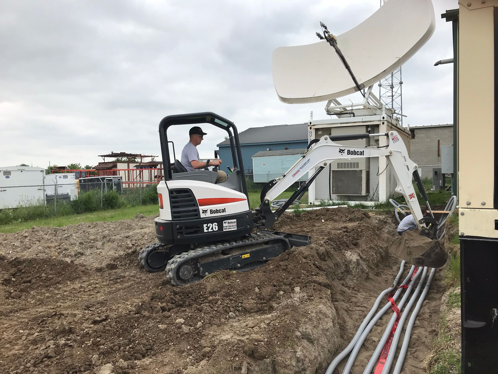 Excavator and satellite dish equipment on a dirt site, with cables laid out in the foreground.