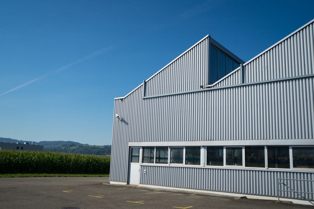 Corrugated metal building with sawtooth roof against a blue sky, fields in background.