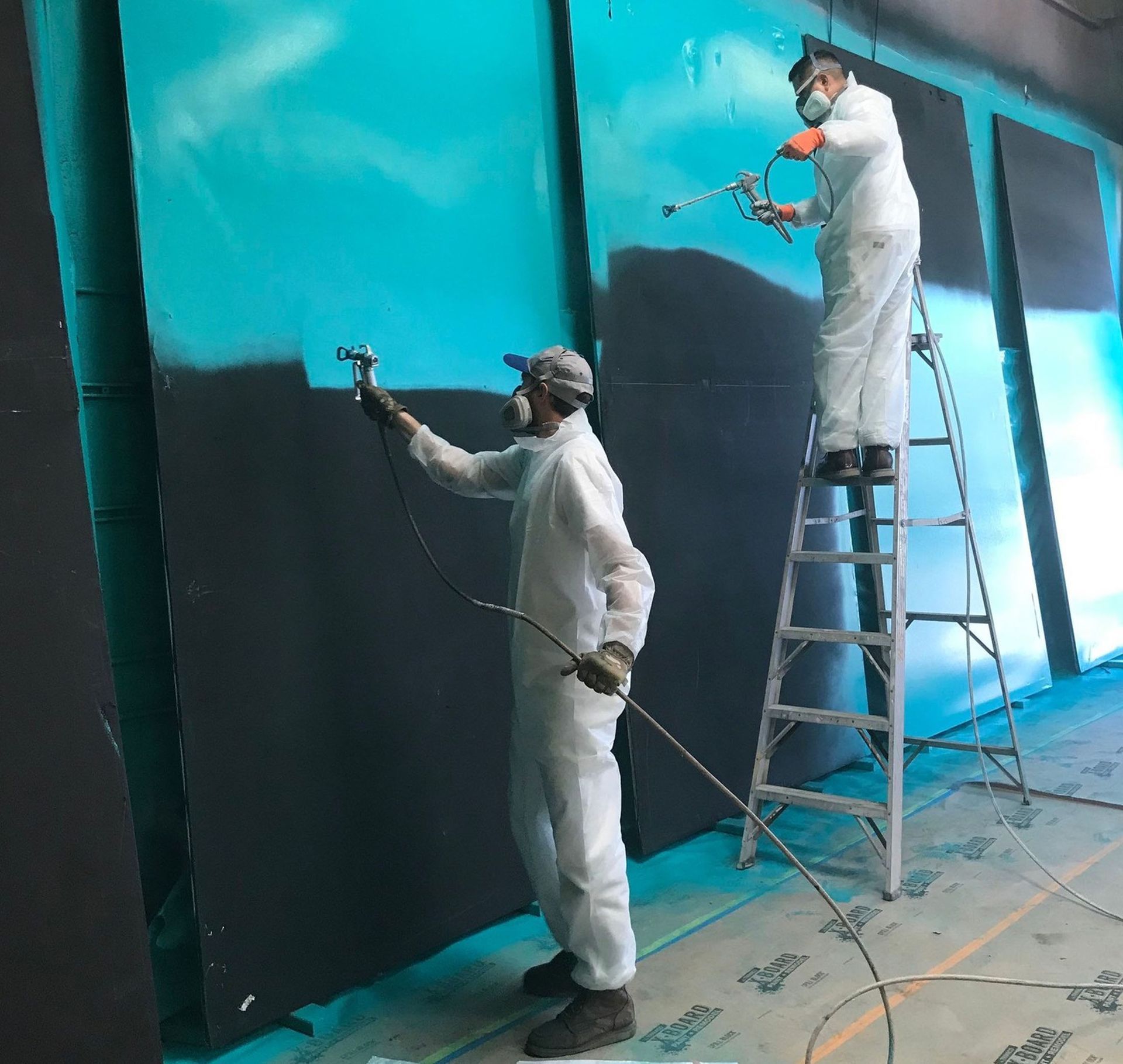 Two people in white protective suits spray painting a dark wall. One on a ladder, in an industrial s