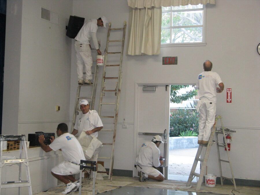 Several painters in white coveralls, using ladders and tools to paint a room with white walls.