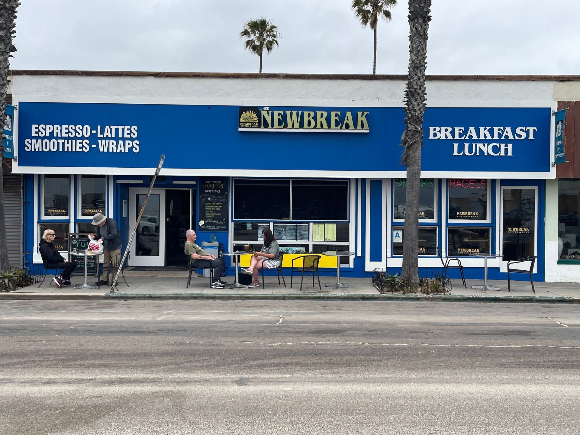 Exterior of Newbreak cafe, blue facade, sidewalk seating, people, palm trees.