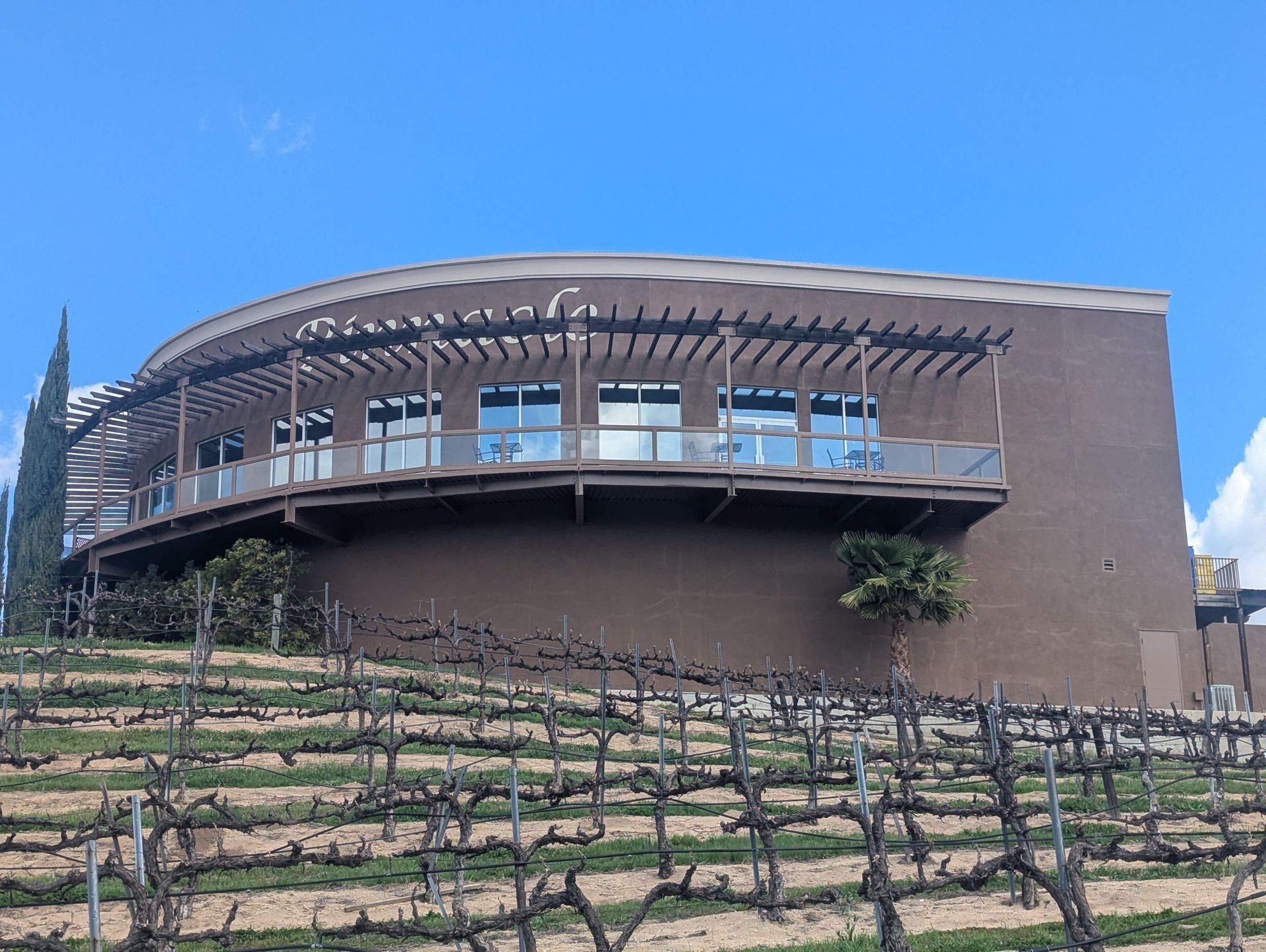 Tan stucco building with arched windows, black iron balconies, and tiled roof against a blue sky.