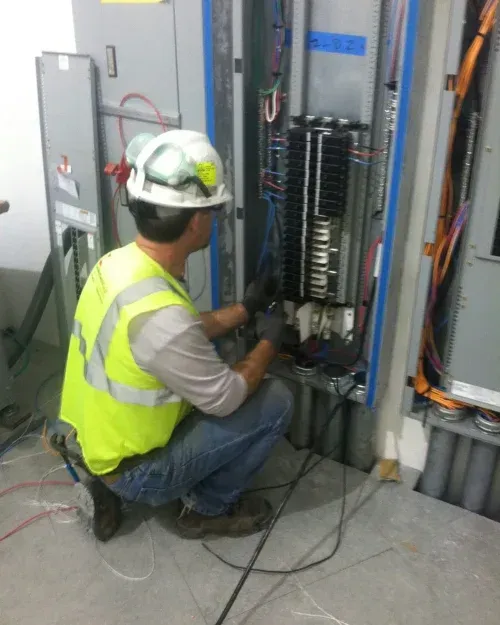 Electrician in a hard hat and safety vest working on a circuit breaker panel in a building.