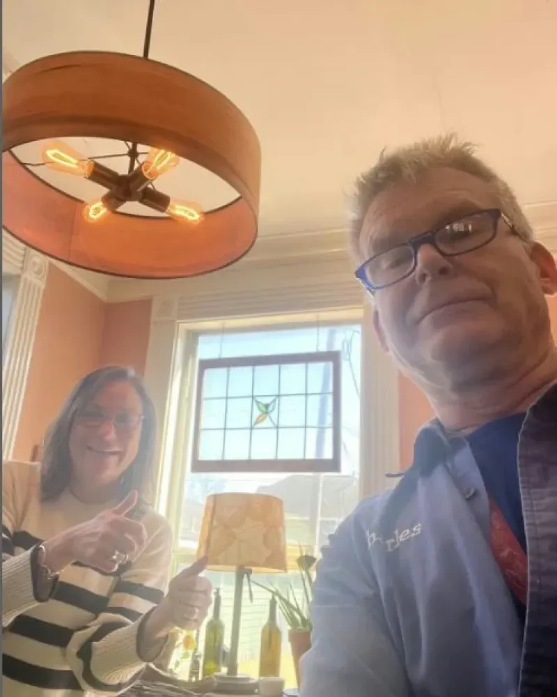 Two people giving thumbs up inside; a man with glasses takes selfie; ornate light fixture.