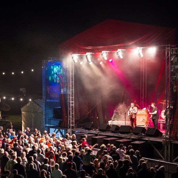 Band performing on stage at outdoor festival, illuminated by spotlights. Crowd watches below.