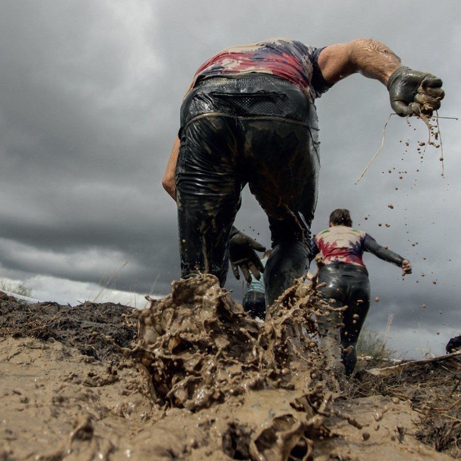 People crawling through thick mud during an obstacle course race under a cloudy sky.