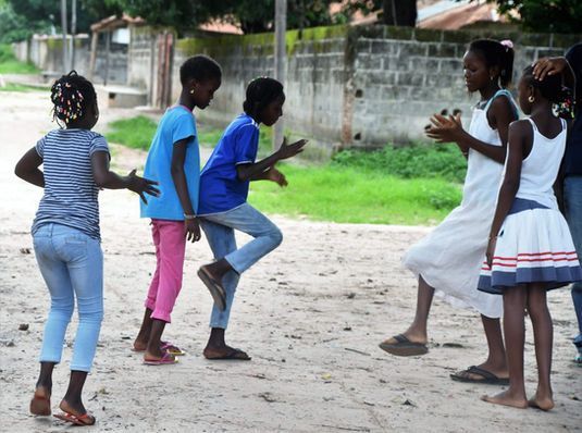 Children playing a clapping game outdoors on a sandy court near a wall and trees.