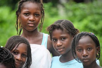 Four children standing outdoors in front of green foliage, looking at the camera
