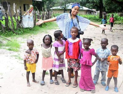 Woman with arms outstretched posing with six children on a dirt path in a village area