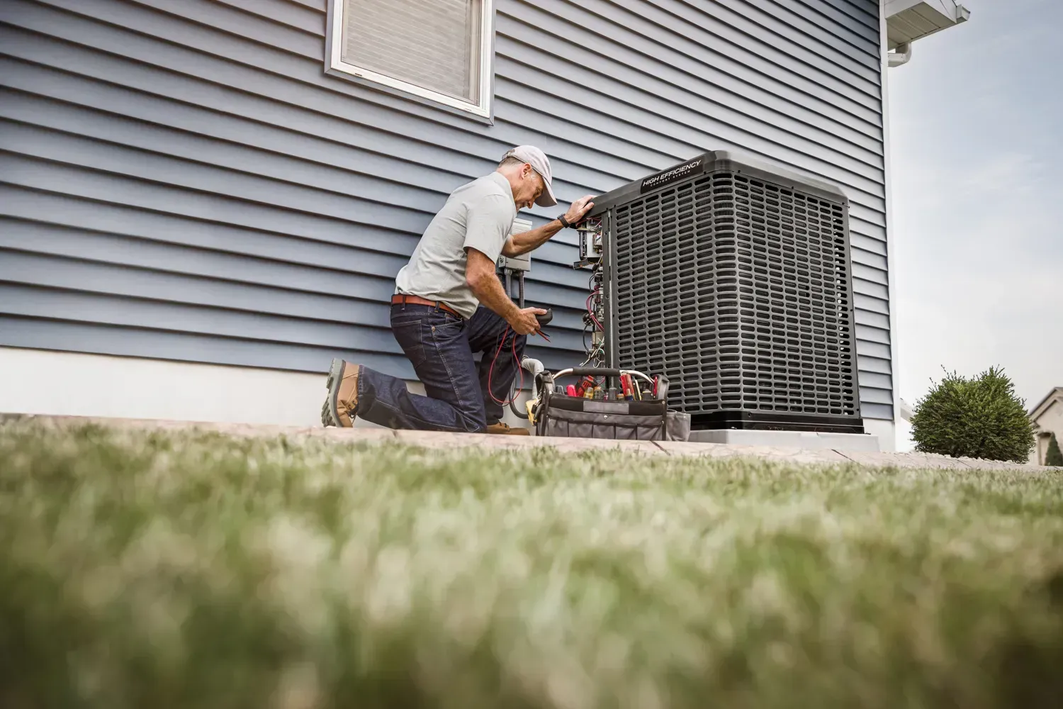 Man kneeling by an outdoor air conditioning unit, working on it. Blue siding, green grass.