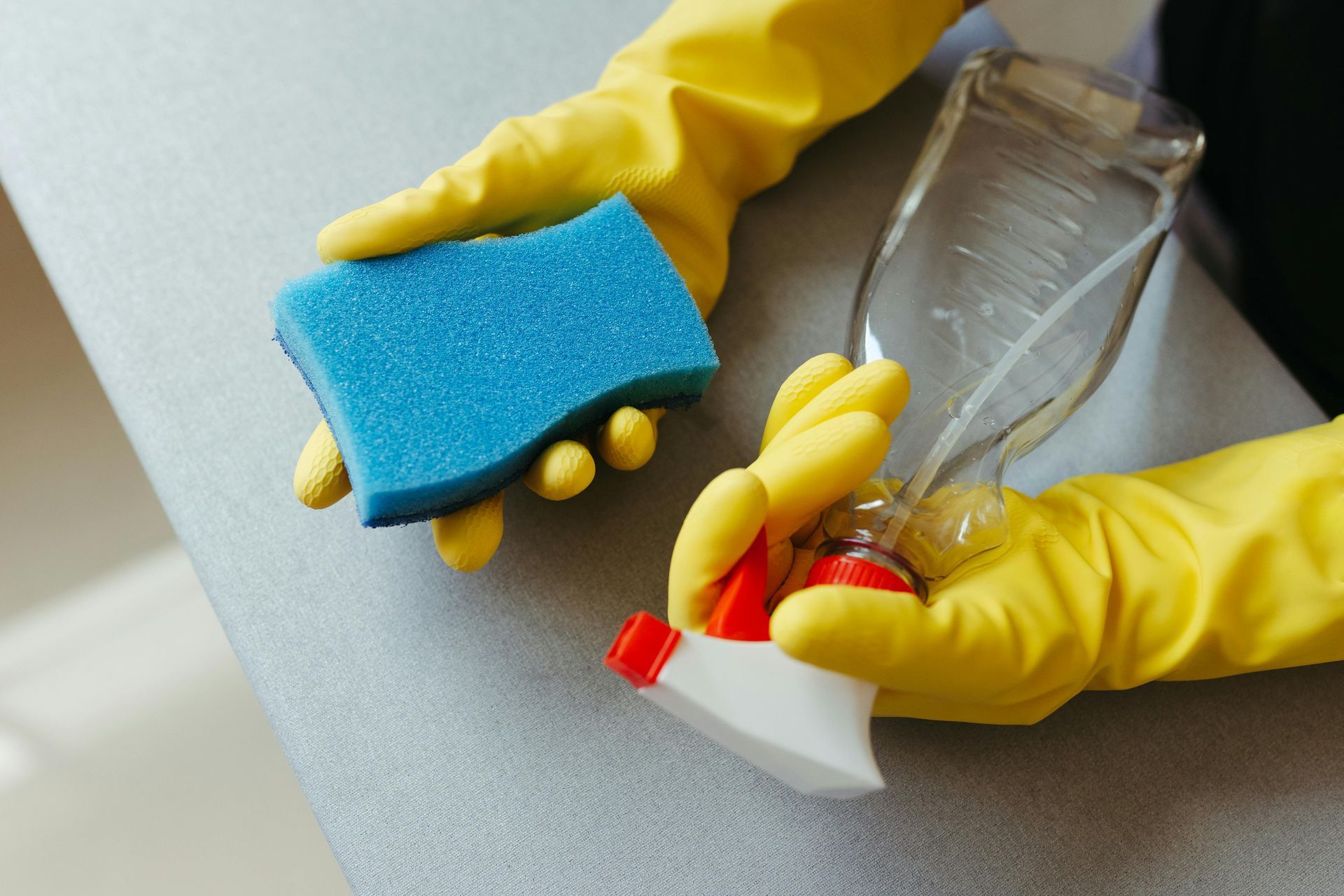 A person wearing yellow gloves is holding a sponge and a spray bottle.