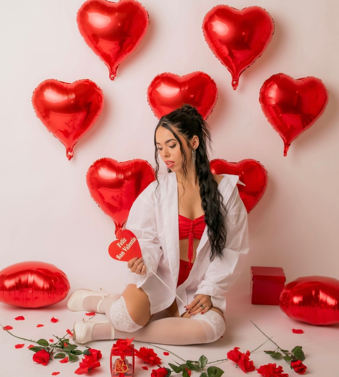 Mujer con lencería roja y bata blanca sosteniendo un corazón entre globos rojos en forma de corazón y rosas en el suelo.