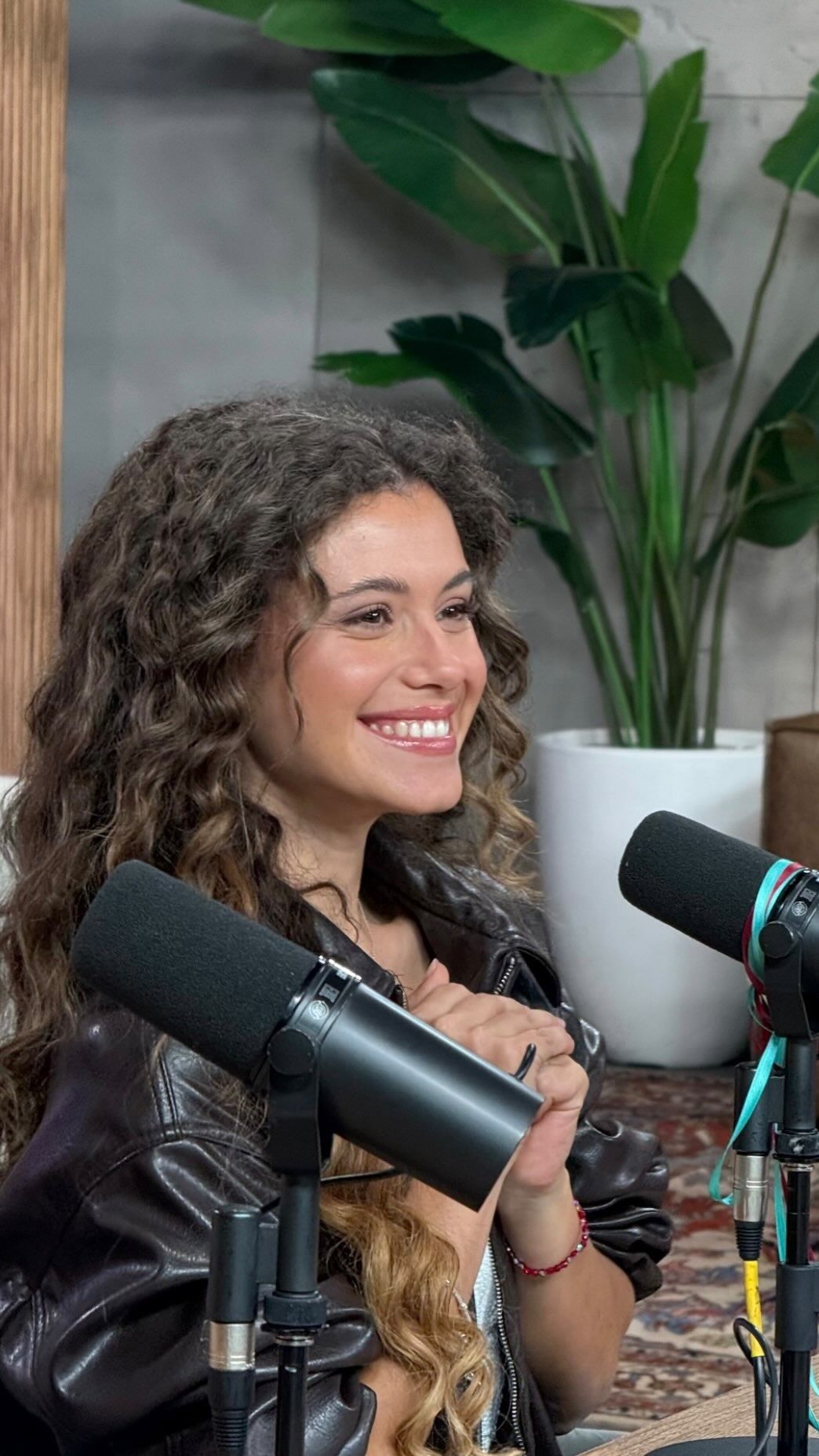 Una mujer sonriente habla ante los micrófonos de un podcast junto a una gran planta frondosa.
