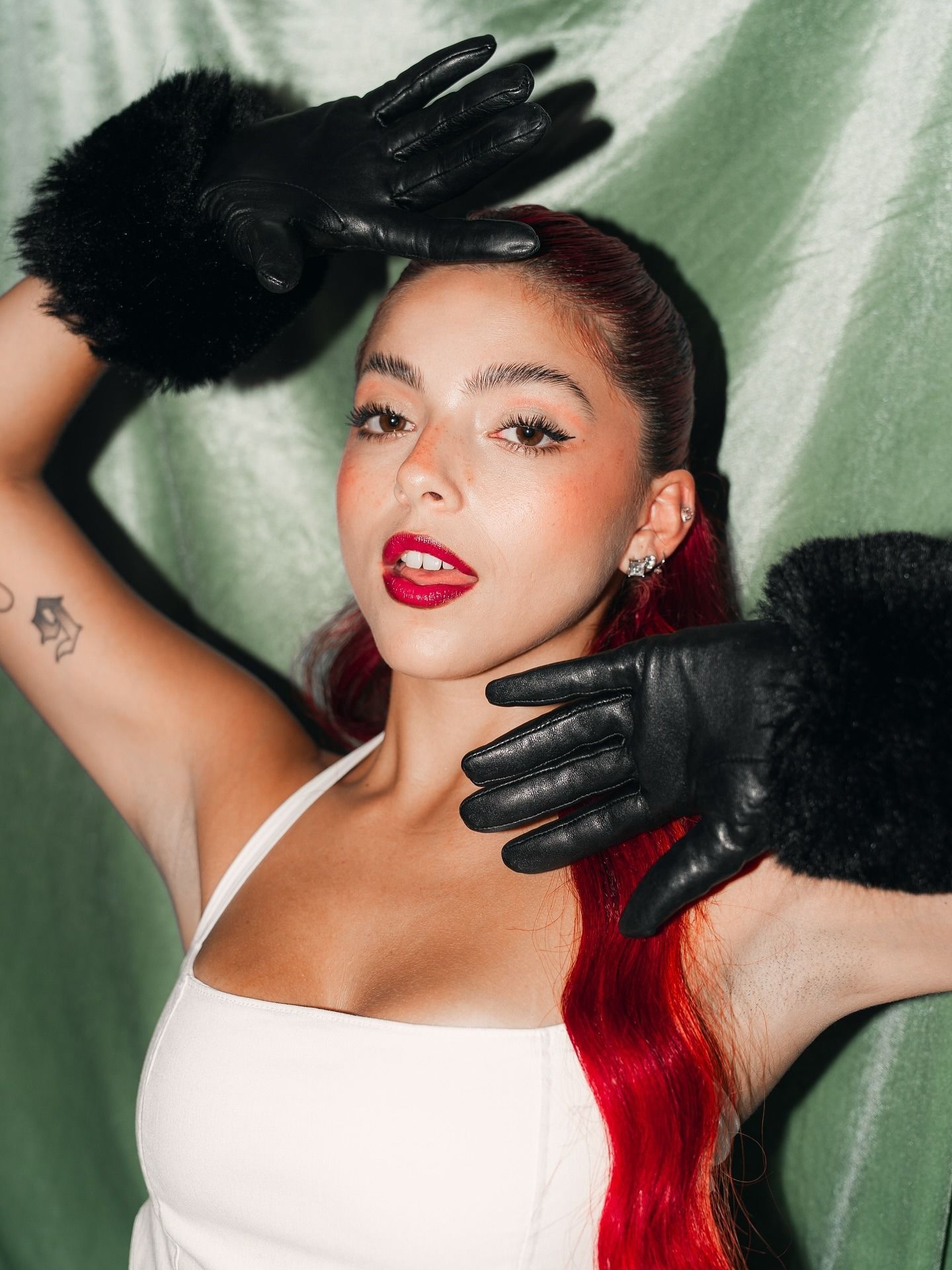 Mujer posando con guantes negros y lápiz labial rojo sobre un fondo verde pálido.