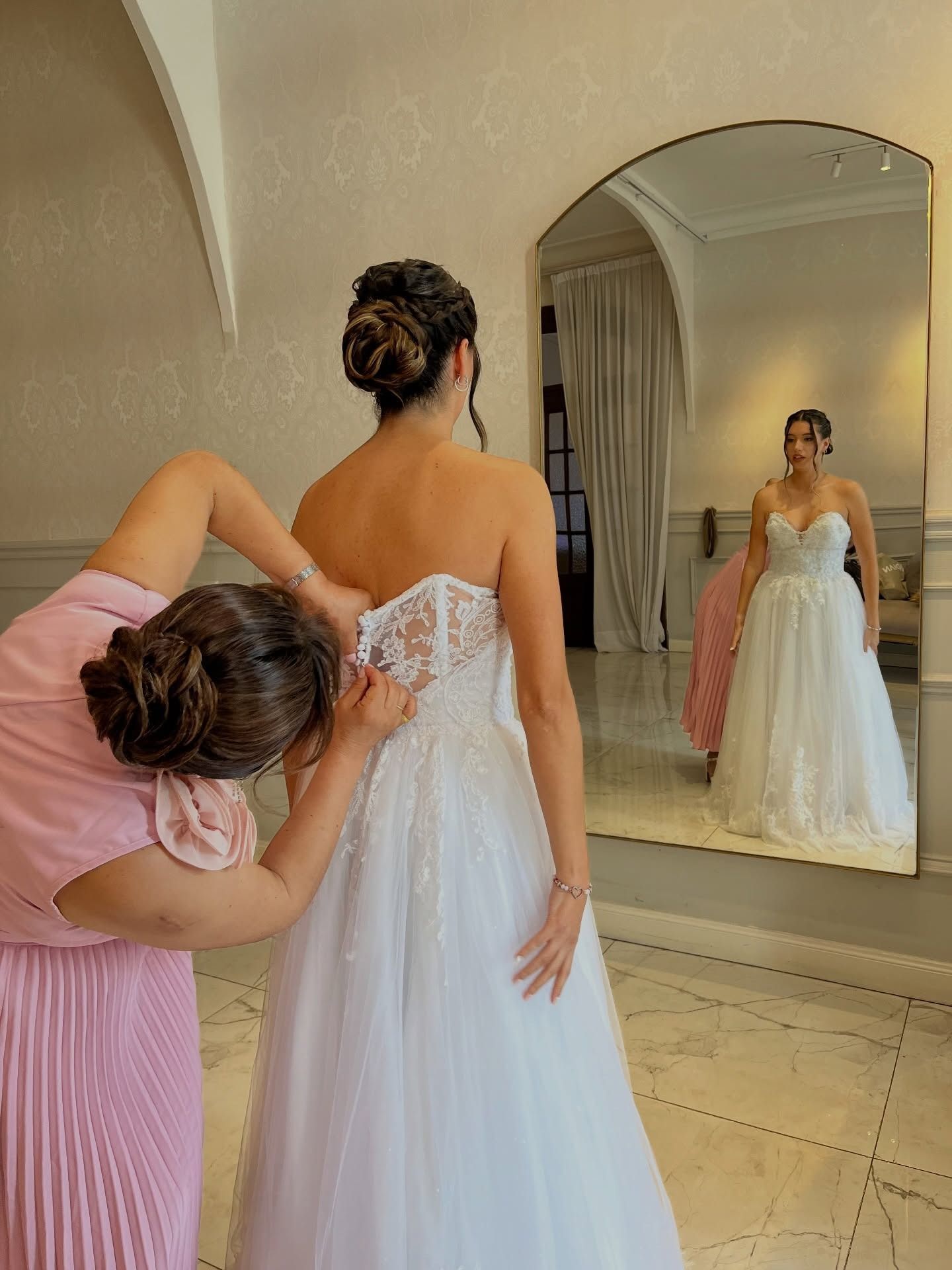Prueba del vestido de novia: una costurera ajusta la espalda mientras la novia se mira en el espejo.