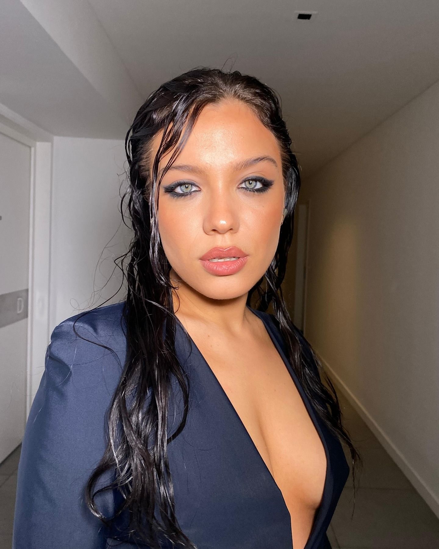 Persona con cabello oscuro mojado y maquillaje de ojos azul, vestida con un blazer azul marino, de pie en un pasillo.