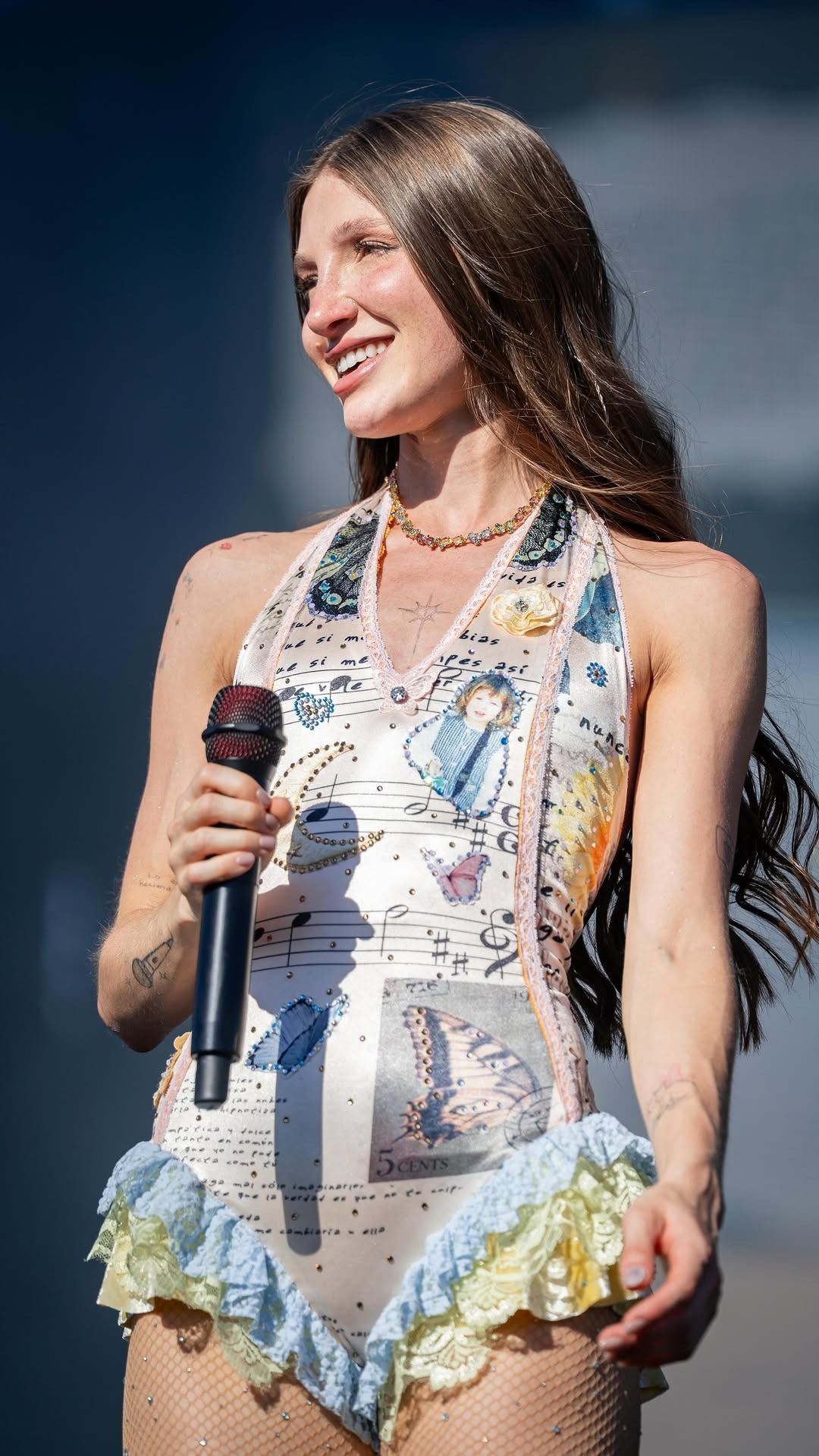Mujer con un vestido blanco estampado sosteniendo un micrófono en el escenario, sonriendo hacia su izquierda.