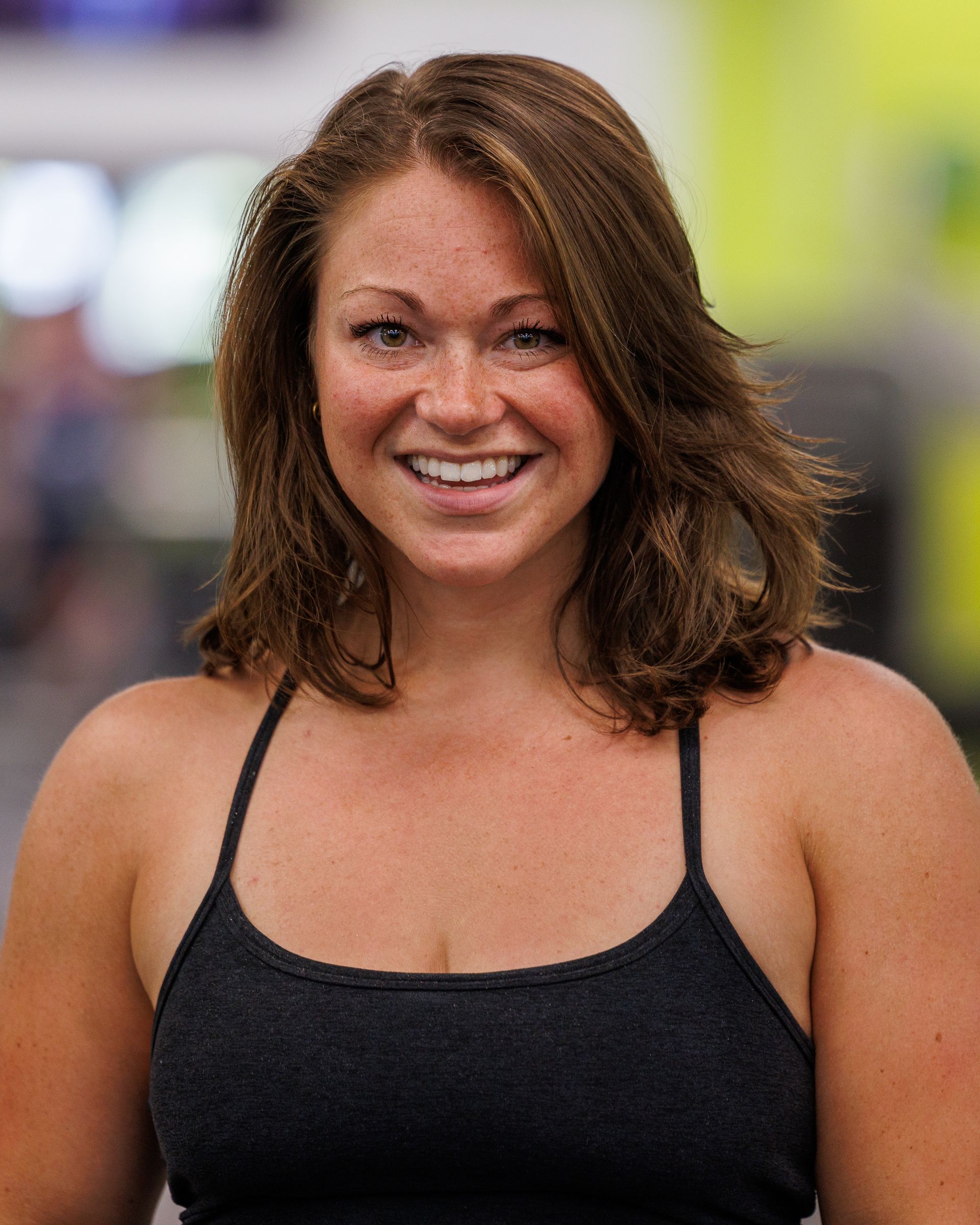 Woman with brown hair smiles, wearing a black tank top, in a gym setting.