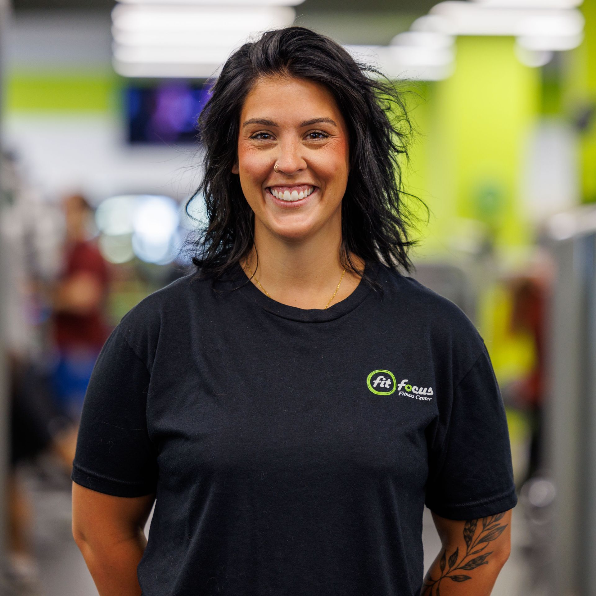 Man with arms crossed, wearing black polo shirt with logo, smiling in gym.