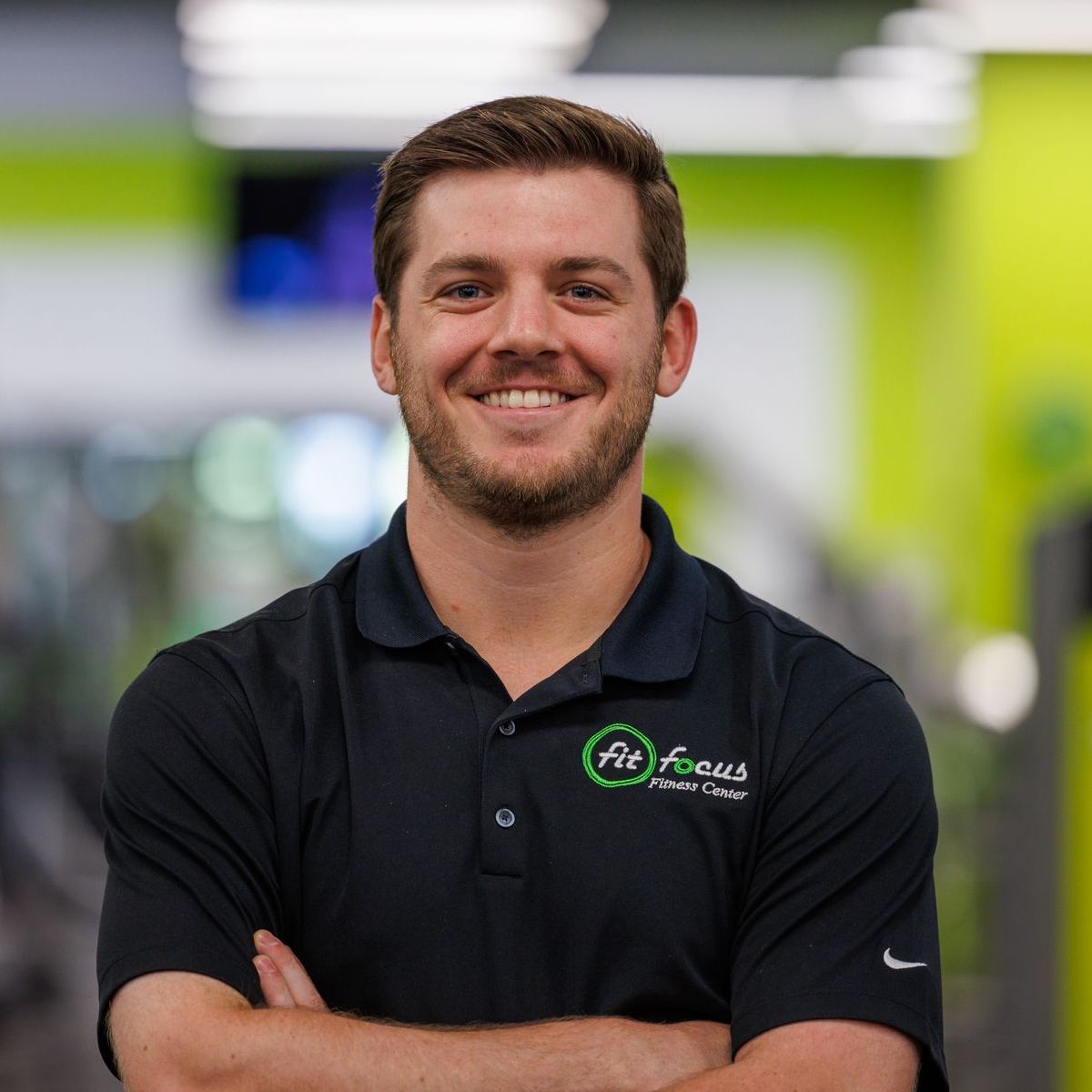 Man with arms crossed, wearing black polo shirt with logo, smiling in gym.