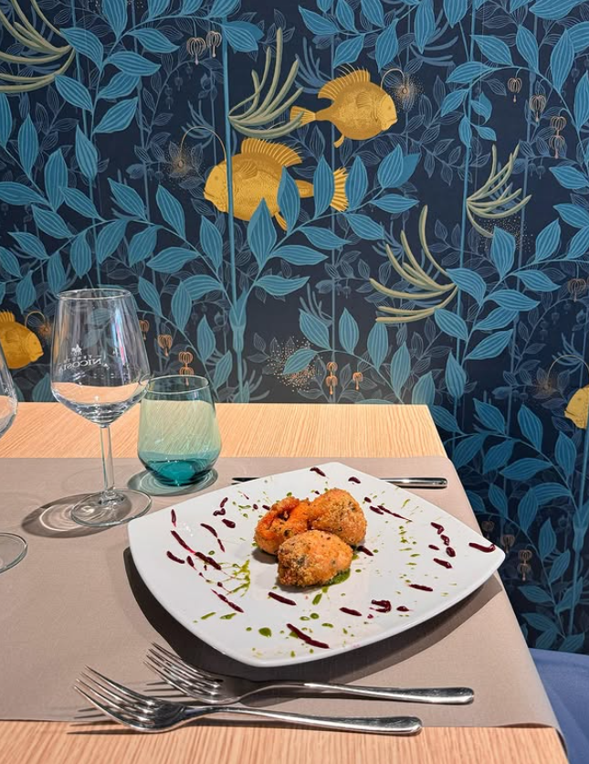 Piatto di cibo su un tavolo apparecchiato per cenare, contro una parete decorata con motivi floreali blu e oro.