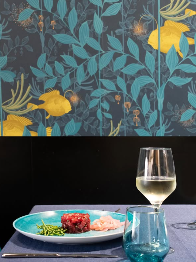 Un piatto con del cibo, un bicchiere di vino e un bicchiere blu su un tavolo con carta da parati a motivi di foglie.