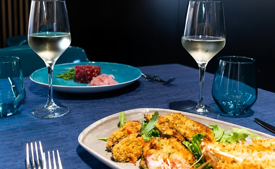 Elegante apparecchiatura da tavola con vino bianco, piatti di pesce e dettagli blu.