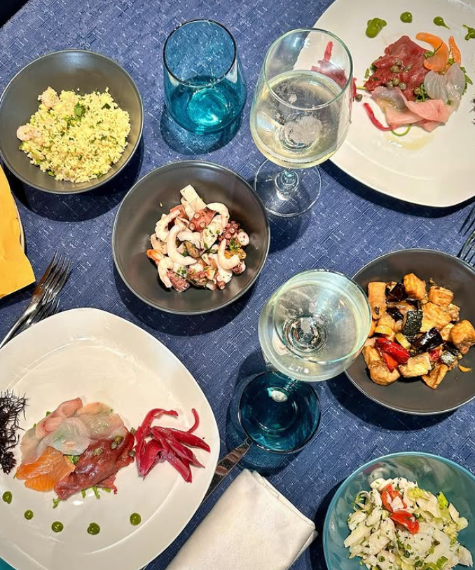 Una tavola apparecchiata per un pasto con piatti di pesce, vino e bicchieri blu su una tovaglia blu.