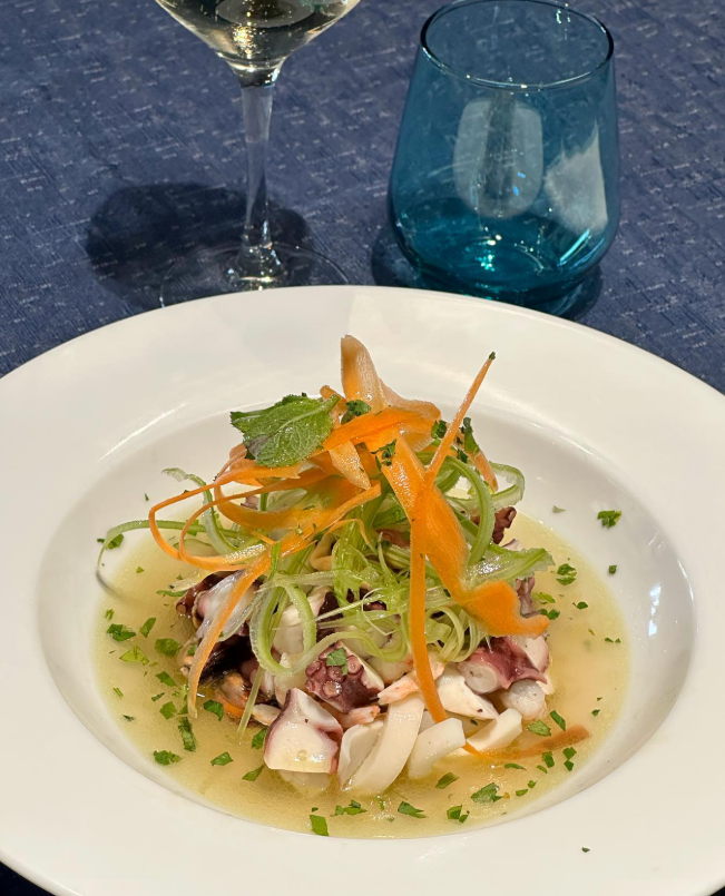 Insalata di polpo in una ciotola bianca, con carote, erbe aromatiche e brodo, più un bicchiere di vino e un bicchiere blu.