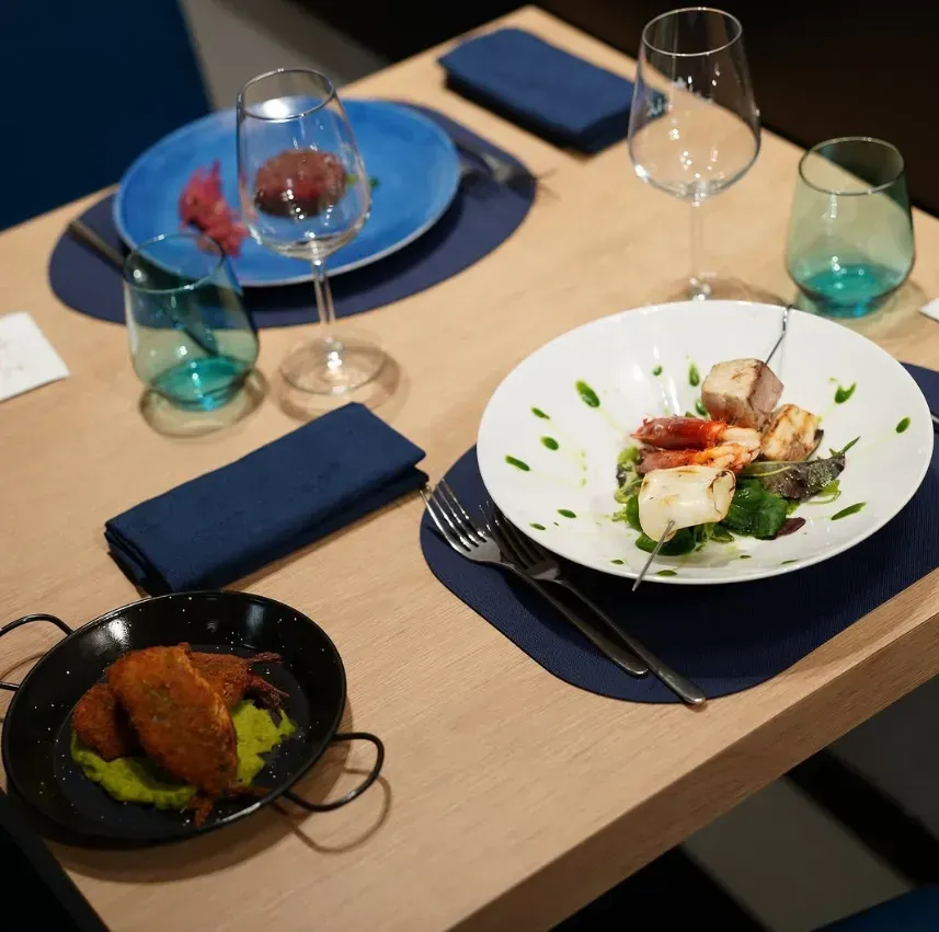 Tavolo del ristorante apparecchiato con piatti, bevande, tovaglioli; piatto bianco con spiedino di cibo.