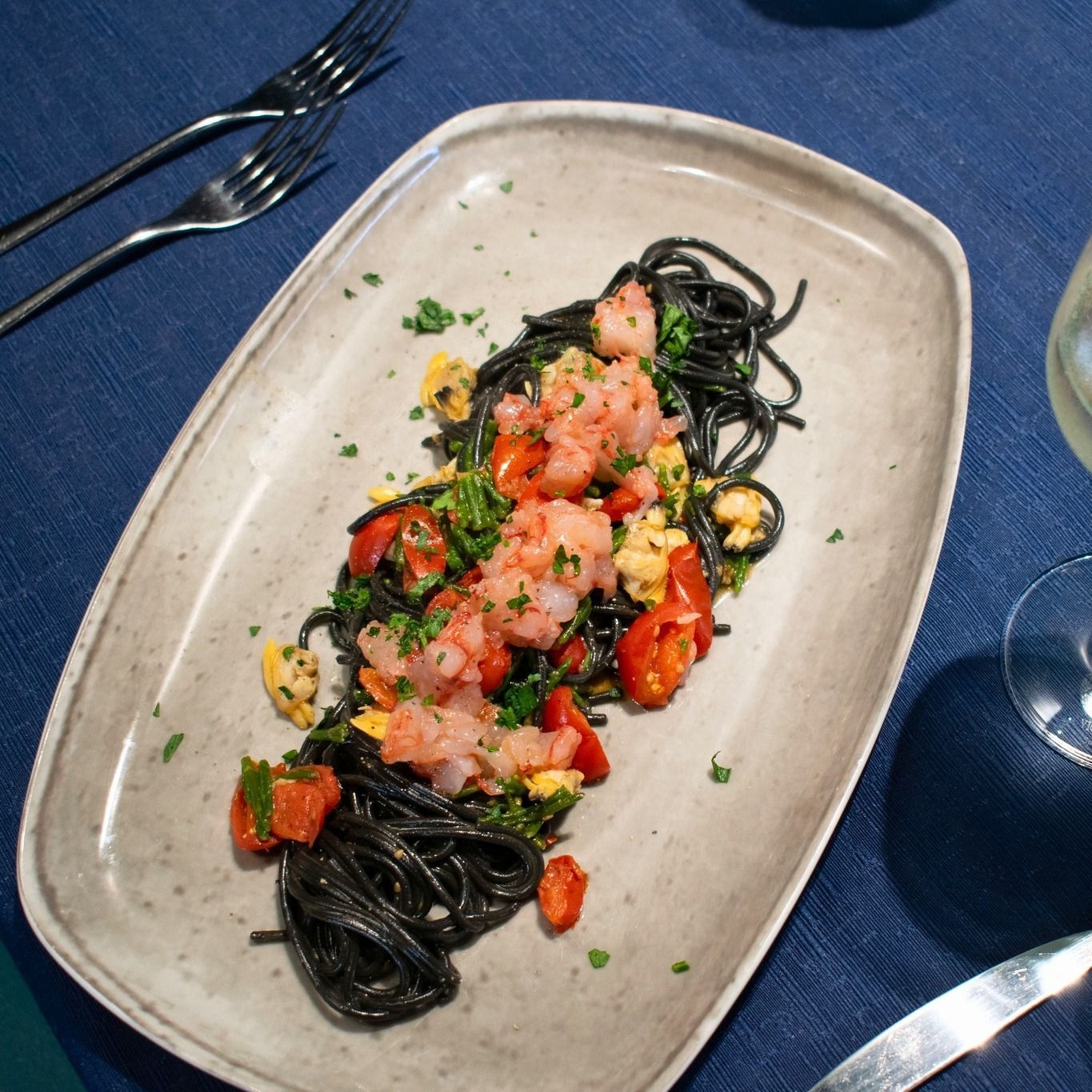 Spaghetti neri con gamberi e pomodori su un piatto rettangolare grigio.