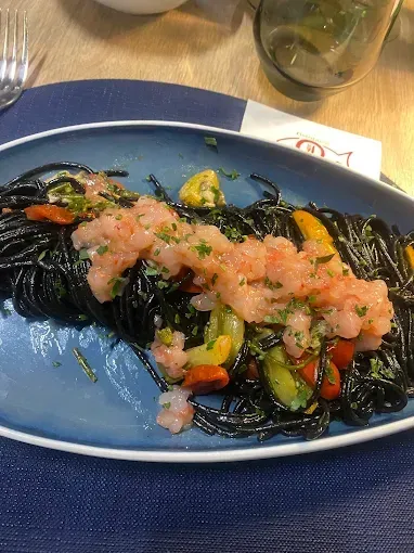 Piatto di spaghetti neri con gamberi rosa e verdure su un piatto blu.