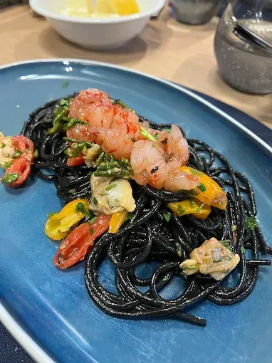 Spaghetti neri con gamberi, vongole e peperoni colorati su un piatto blu.