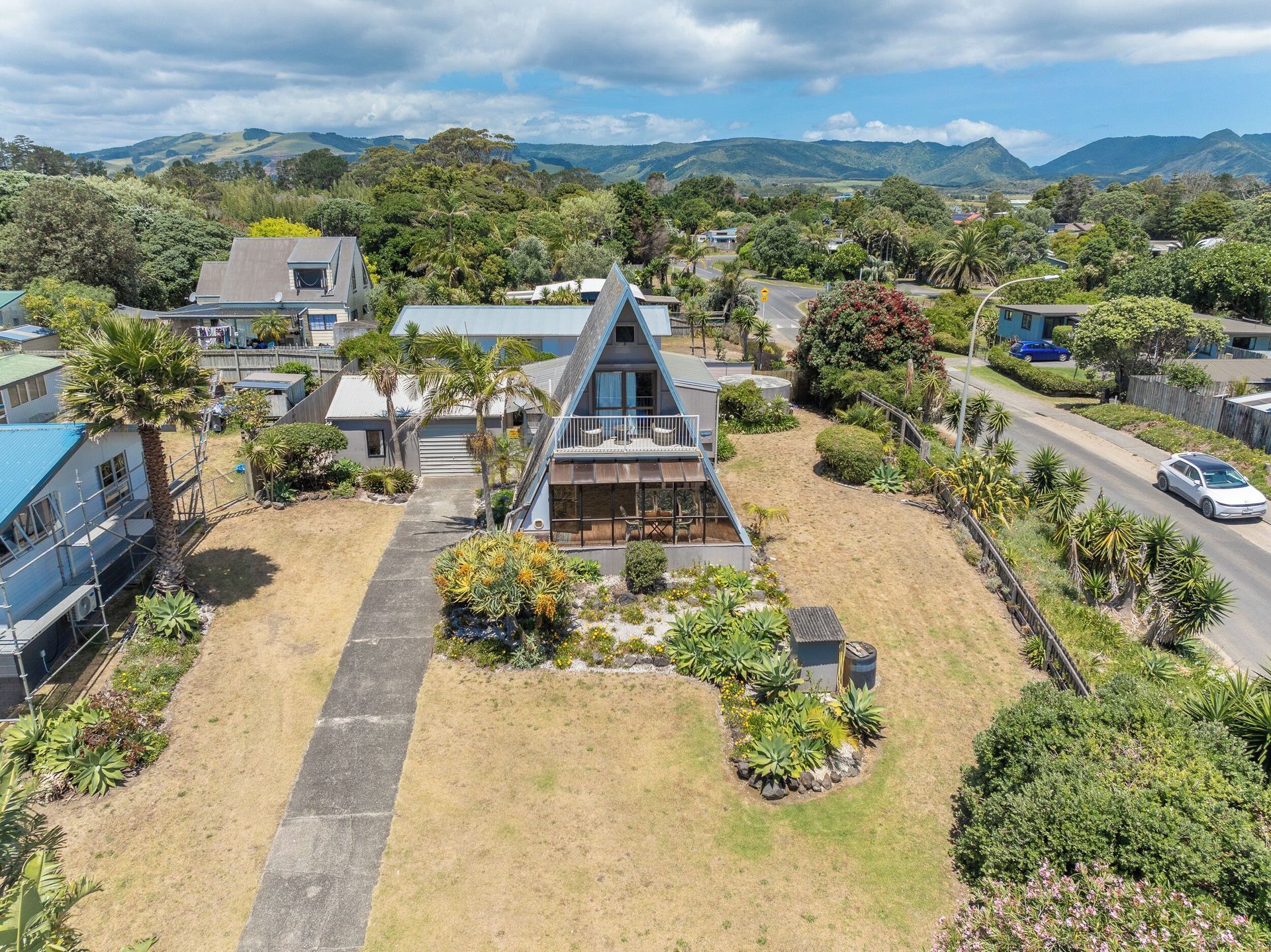 Aerial view of an A-frame house with a balcony, surrounded by greenery and a road.