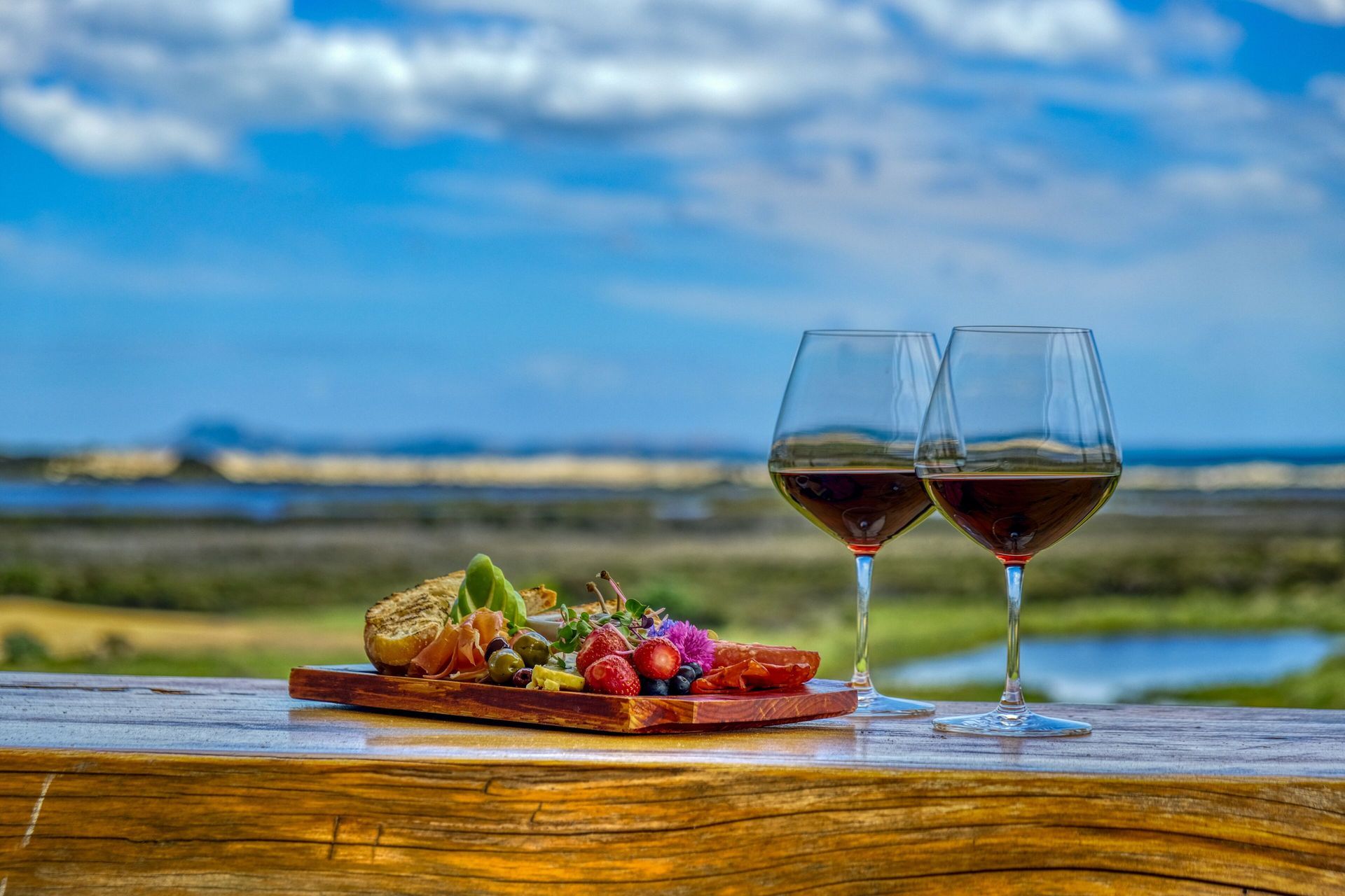 Two wine glasses and a charcuterie board on a wooden table overlooking a landscape with blue skies.