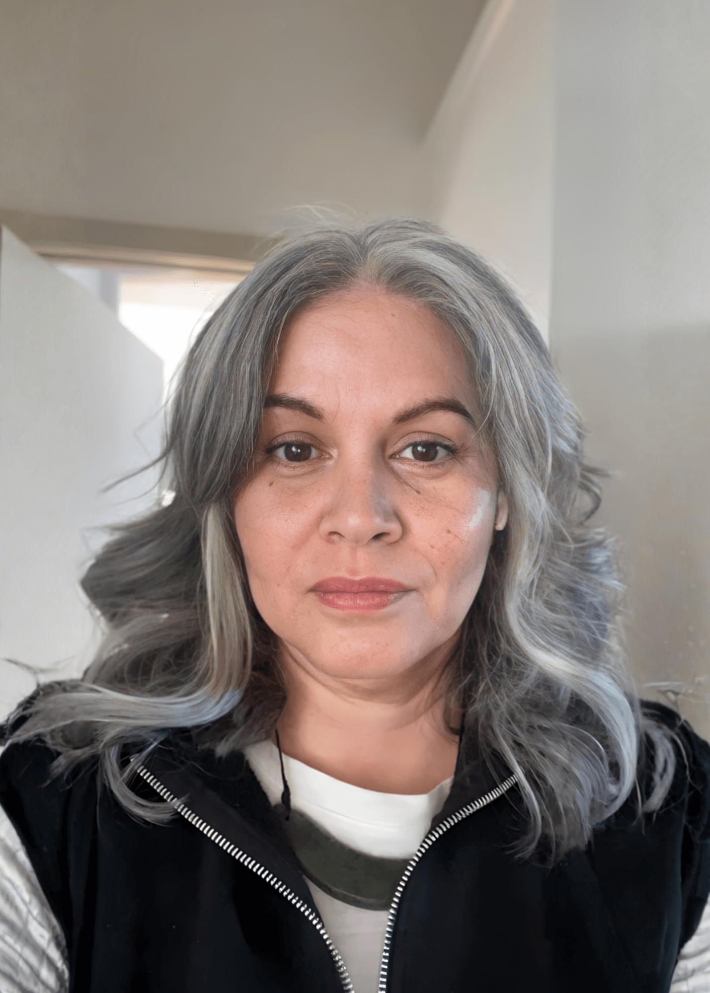 Woman with gray wavy hair, wearing a black vest and white shirt, in a hallway.