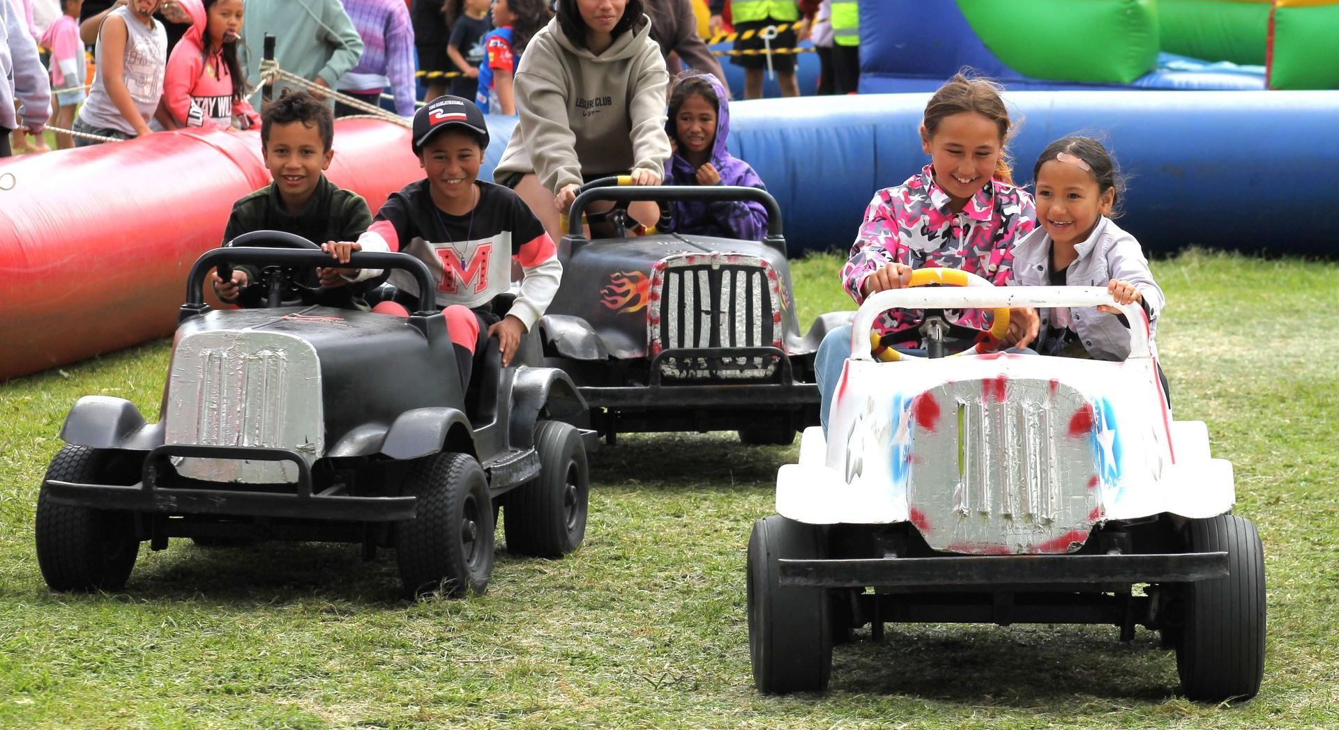 Children in toy cars, smiling, outdoors on grass, near an inflatable slide.