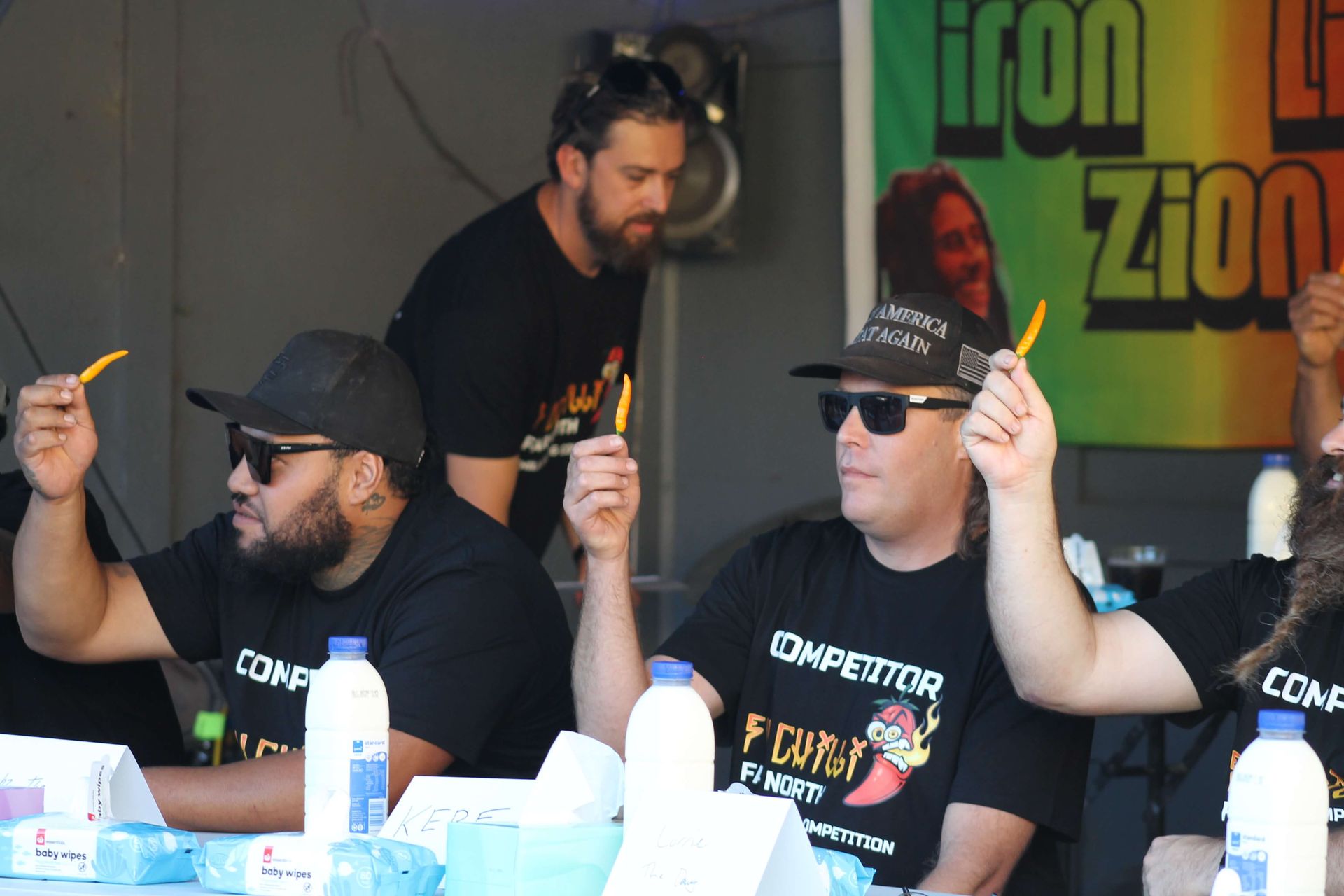 A group of men are sitting at a table at chilli comp in front of a sign that says iron zion