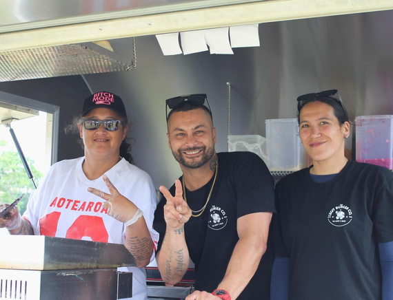 Three people smiling at a food truck: One makes a peace sign, one a victory sign, and all wear black shirts.
