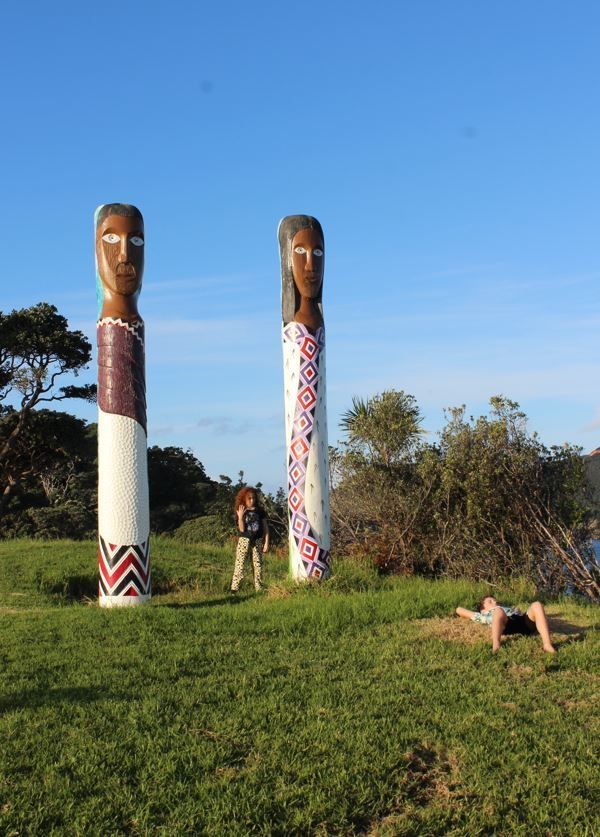 Two tall, decorated Māori carvings with people nearby on a grassy area, under a blue sky.
