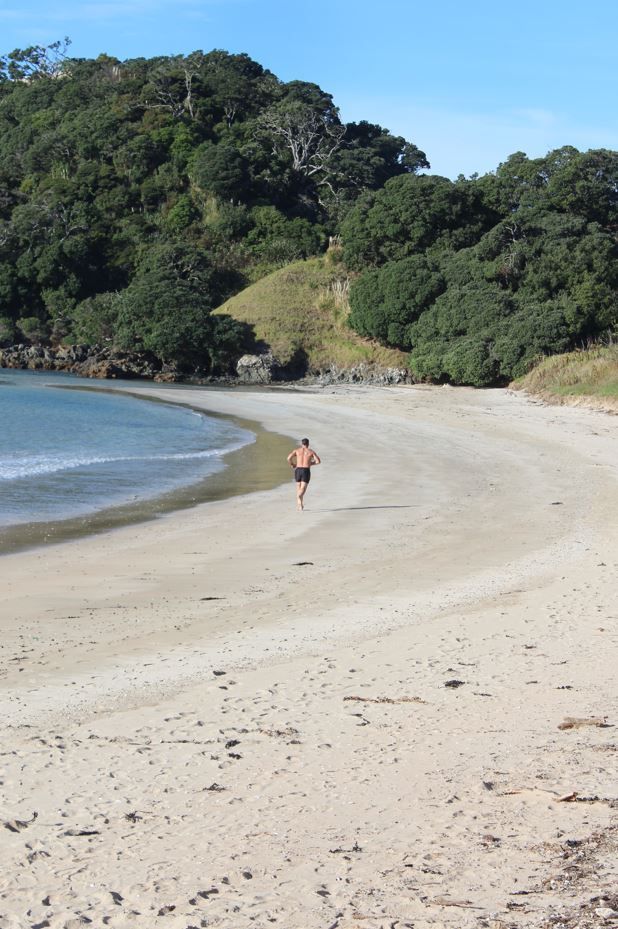 Man running on sandy beach, ocean and lush green hills in background.