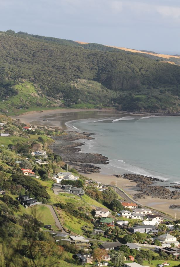 Coastal town with houses on a hillside, overlooking a beach, bay, and forested hills.