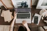Person typing on a laptop at a desk with books, phone, and picture frames.