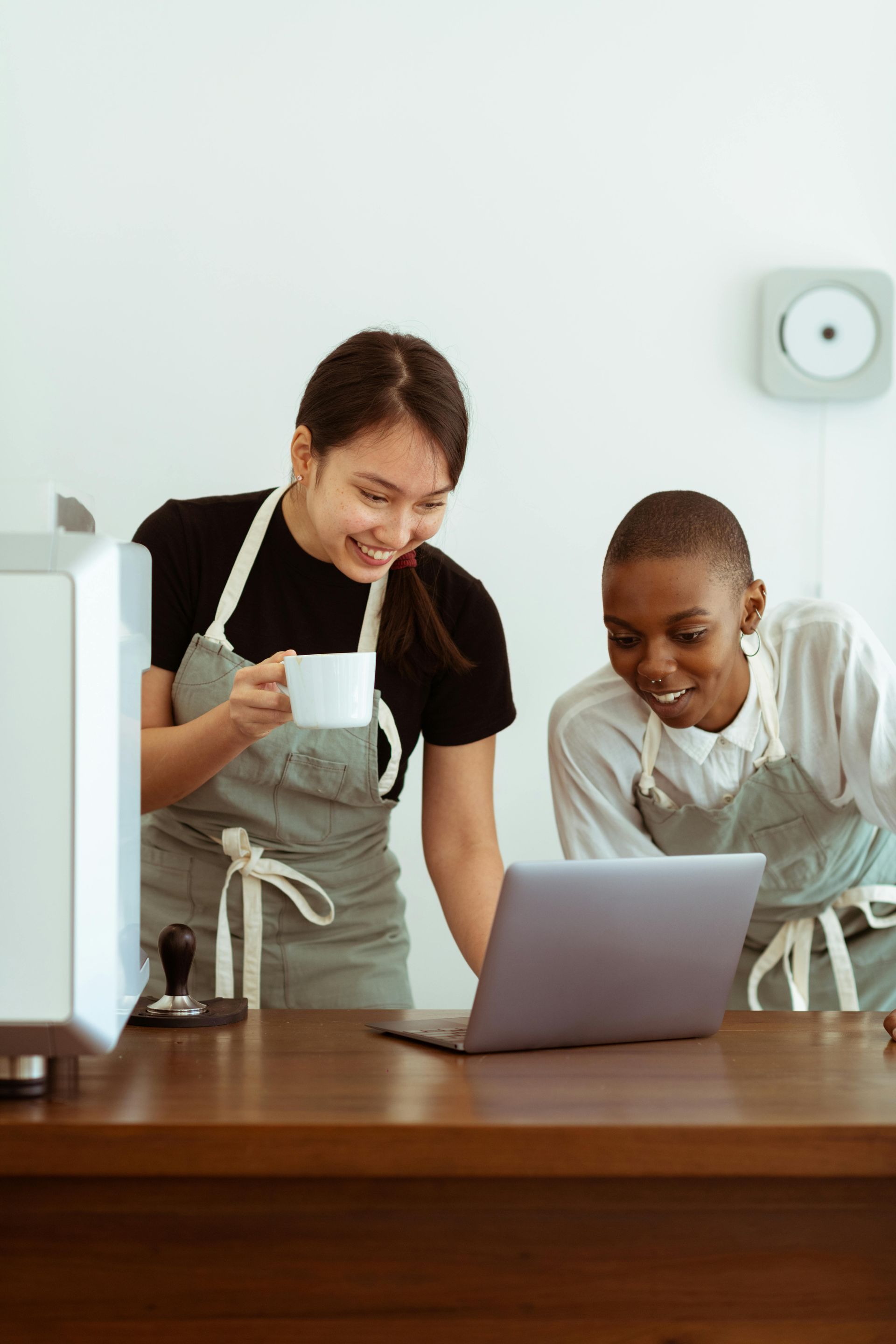 Two baristas laugh while looking at a laptop at a coffee shop counter.