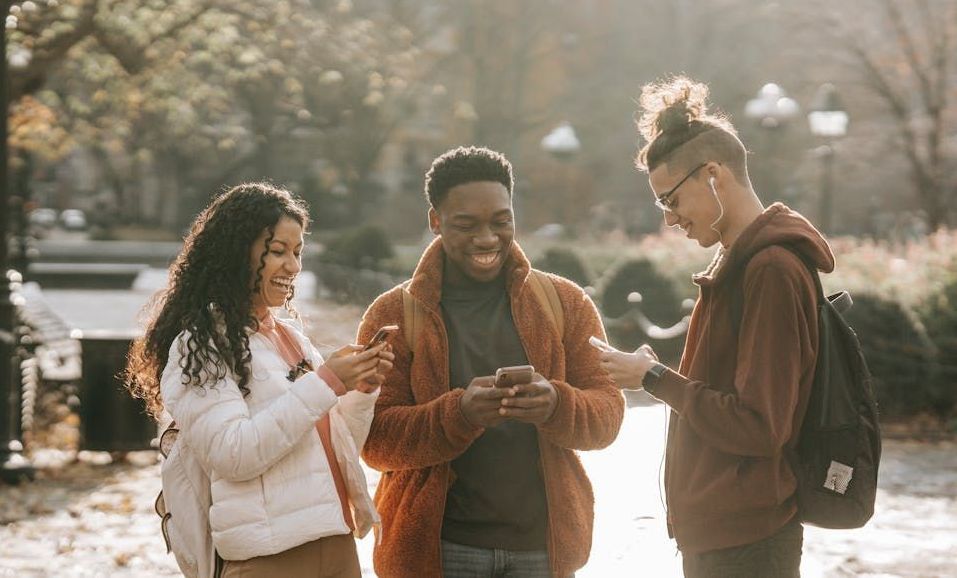 A group of young people are standing in a park looking at their phones.