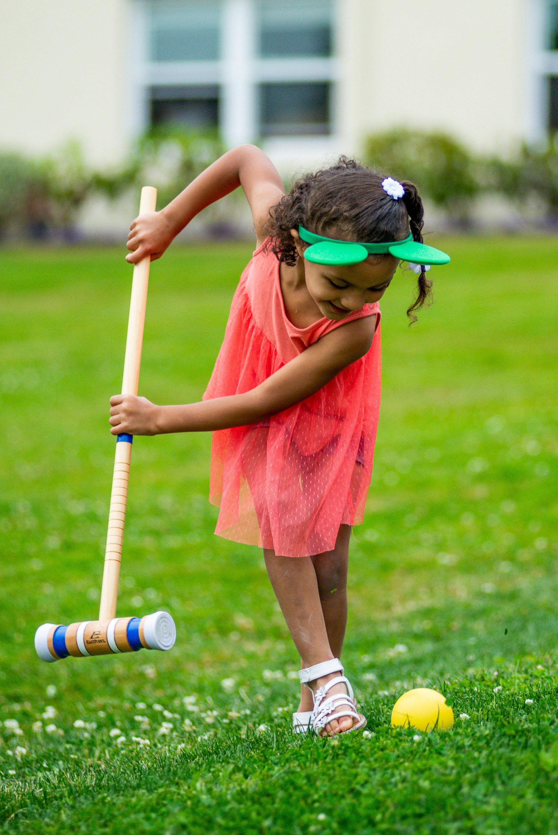 Girl in red dress playing croquet on a green lawn, smiling while hitting the ball with a mallet.