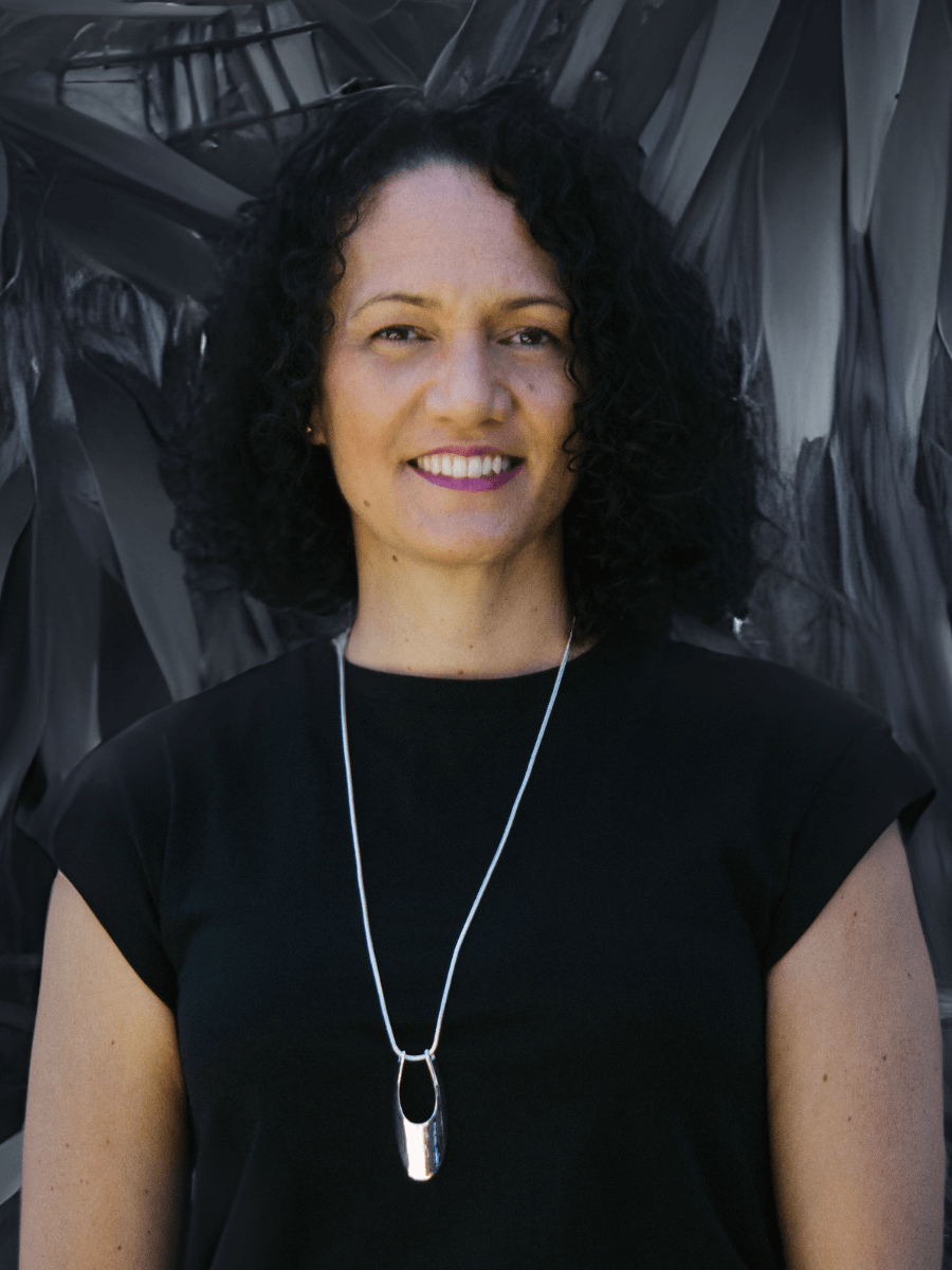 Woman with curly dark hair smiles, wearing a black top and silver necklace; blurred leafy background.