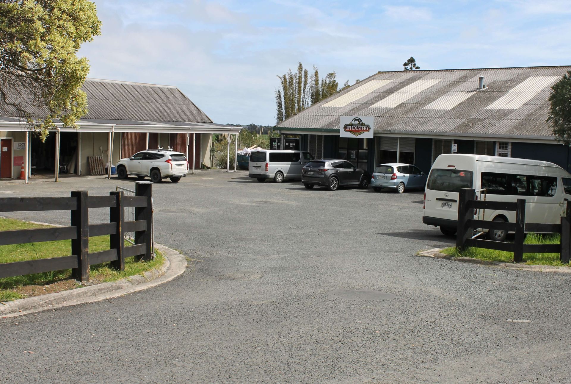 Gravel driveway leading to buildings. Cars and vans parked outside a shop with a sign. Fence in foreground.