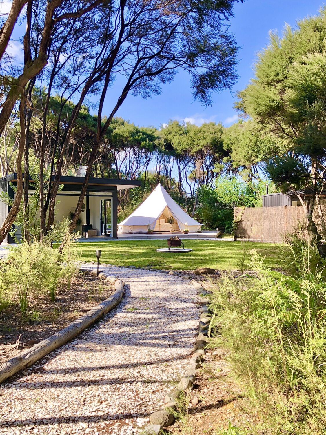 Path leads to white canvas tent and small modern building in a green, wooded area.