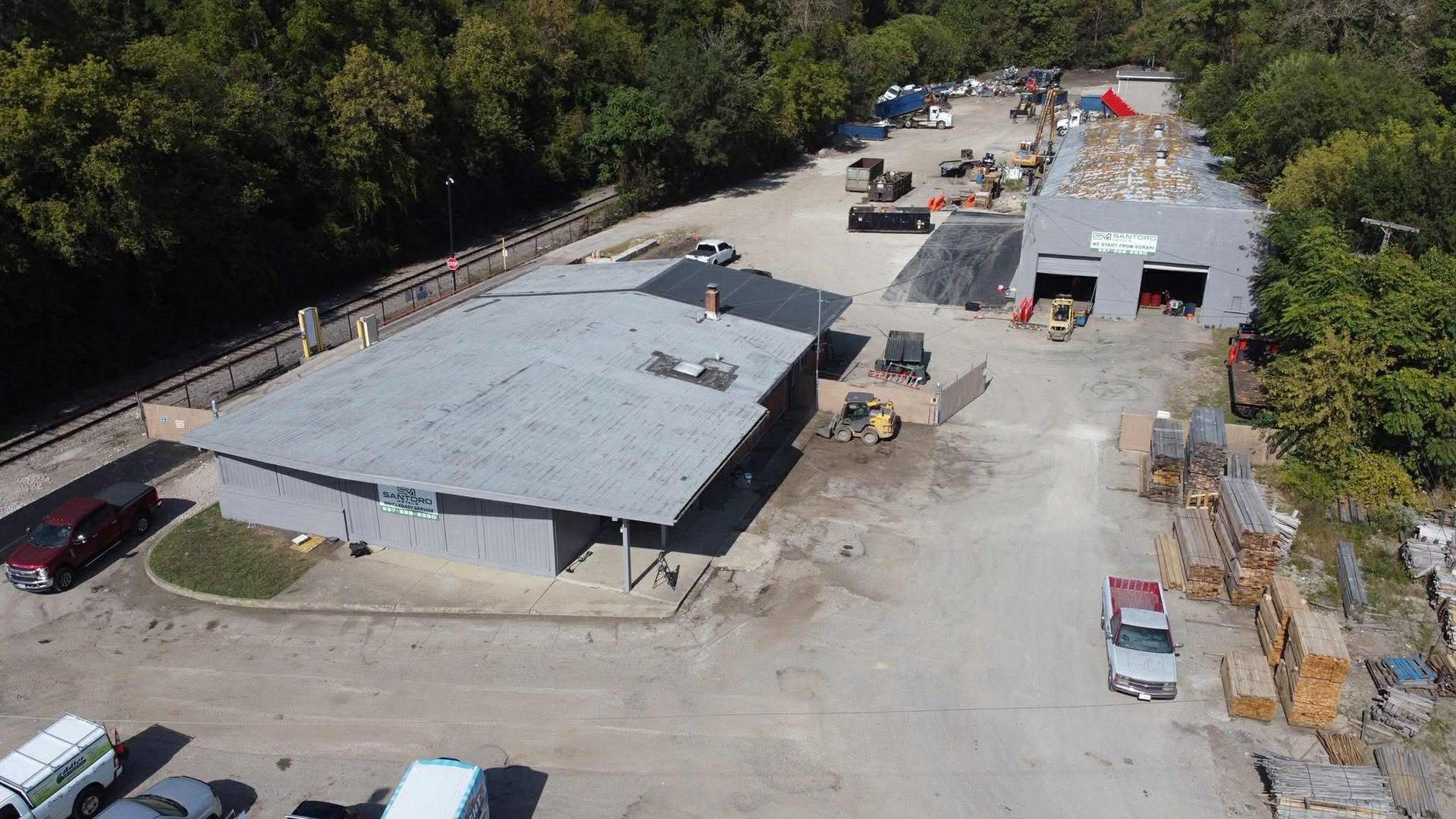 Aerial view of a gray building and surrounding lot with vehicles, trees, and construction materials.
